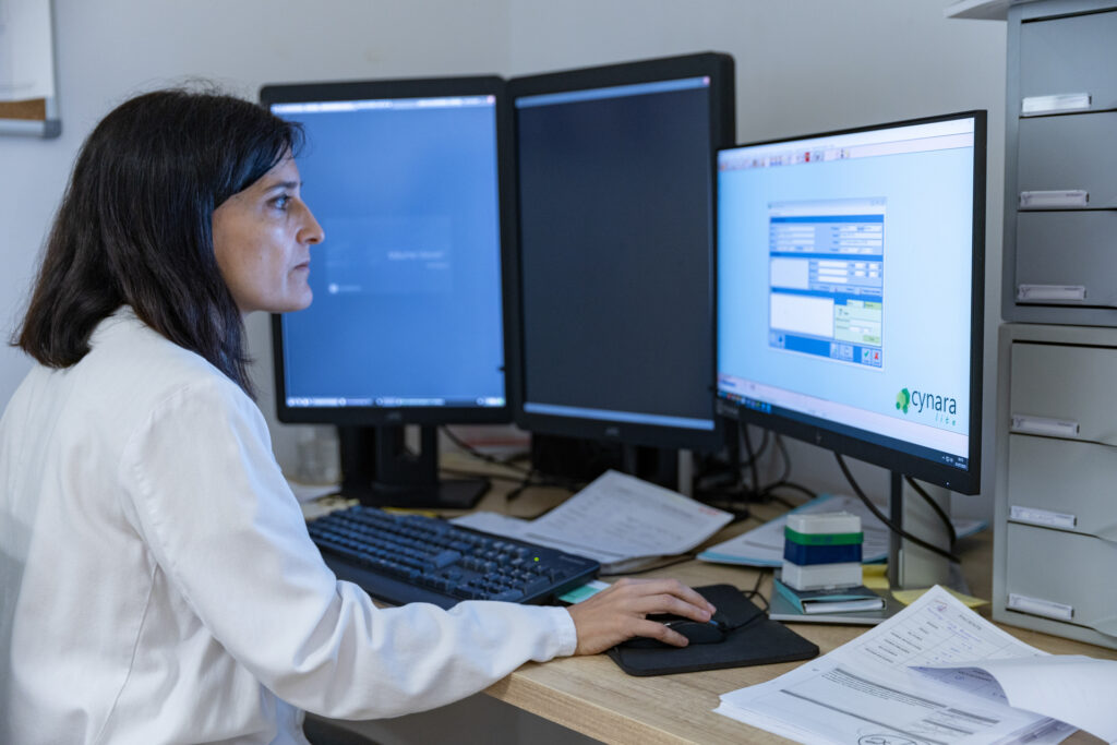 Person working in an office in front of three monitors, using a keyboard and mouse, with documents and office supplies on the desk.